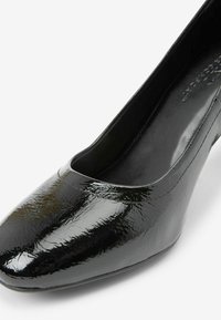 Black patent leather pump with a rounded toe and slightly textured surface. Features a sleek silhouette and flexible design.