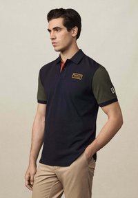 Námornícky modrá polo košeľa vyrobená z bavlny, s krátkymi olivovými rukávmi, červeným golierom a nášivkou loga na hrudi.