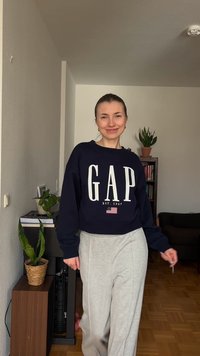 Νεαρή γυναίκα που χαμογελά, φοράει ένα μπλε GAP sweatshirt και γκρι φόρμα, στέκεται σε ένα σαλόνι με φυτά και μια βιβλιοθήκη στο φόντο.