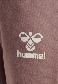 Hummel-logo met een symmetrisch ontwerp in het wit op een textuurrijke, gedempte paarse stofachtergrond, met een strakke lettertype onder het logo.