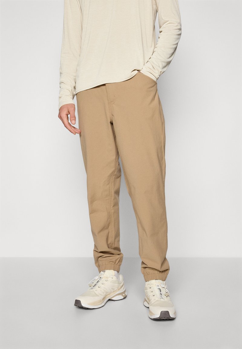 Man in beige jogger pants and cream long-sleeve shirt with one hand in pocket and wearing cream and white sneakers, standing against plain background.