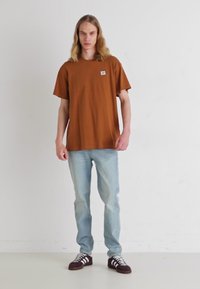 Camiseta marrón de manga corta con un pequeño logo, combinada con jeans azul claro y zapatos oscuros con rayas blancas. Conjunto casual sencillo.