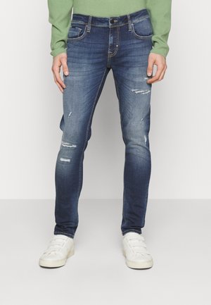 Mann trägt schlanke blaue abgenutzte Jeans, weiße Sneakers und ein hellgrünes Langarmshirt, steht vor einem einfarbigen Hintergrund.