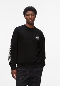 Junger Mann trägt einen schwarzen Pullover mit "Karl Lagerfeld"-Schriftzug auf dem Ärmel und einem kleinen Grafikdesign auf der Brust, kombiniert mit dunkel gemusterten Hosen.