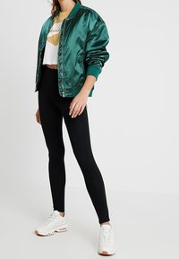 Grön satin bomberjacka, kort vit t-shirt med grafik, svarta leggings med sidosträck, och vita träningsskor med beige sula.