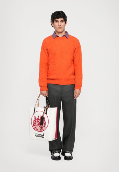 Orange textured sweater, gray trousers, black and white shoes, holding a white tote with red graphic details and leather straps.
