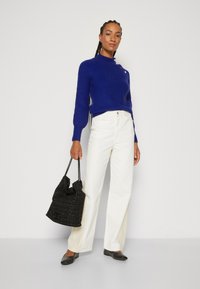 Blue knitted sweater with round neckline and accent buttons, white wide-leg pants, black textured bucket bag, and black flats.