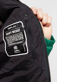 Schwarze schwere Jacke mit grünem Innenfutter; ausgestattet mit einem Reißverschluss und einem großen weißen und schwarzen Pflegeetikett mit Informationen zum Wärmeschutzlevel.