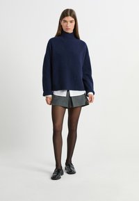 Granatowy sweter z golfem o luźnym kroju, z prążkowanymi mankietami, połączony z szarym, plisowanym mini spódnicą i czarnymi butami, noszony na przezroczystych czarnych rajstopach.