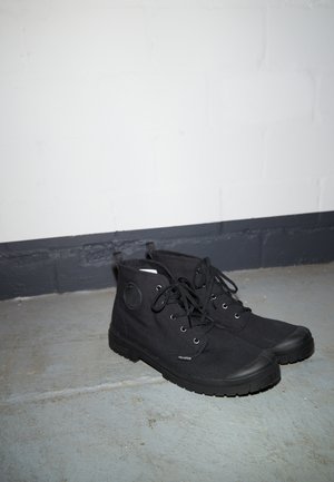 Schwarze hohe Canvas-Stiefel mit Schnürsenkeln, Gummi-Zehenschutzkappen und robusten Sohlen, die auf einem grauen Betonboden nahe einer weißen Wand stehen.