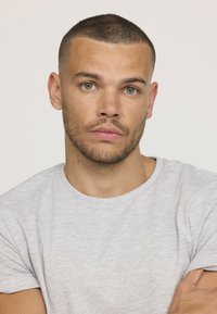 Homme aux cheveux courts portant un t-shirt gris clair à col rond. Il a une légère barbe et fait face à l'appareil photo les bras croisés.