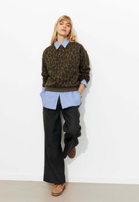 Pull vert olive à imprimé léopard superposé à une chemise boutonnée bleu clair, assorti à un pantalon gris foncé à jambes larges et des chaussures marron.