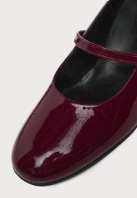 Chaussure Mary Jane en cuir verni rouge foncé brillant, à bout arrondi et bride unique sur semelle intérieure noire sur fond blanc.