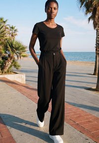 Woman in black t-shirt and wide black pants walking on a seaside promenade with palm trees and ocean in the background.
