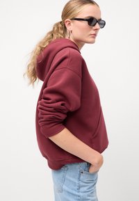 Maroon dukserica s kapuljačom izrađena od mekog materijala, s kengurskim džepom, rebrastim manžetama i rubom; uparena sa svijetloplavim farmerkama.