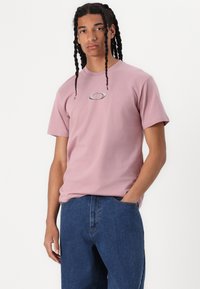 T-shirt de algodão cor-de-rosa com mangas curtas e um logótipo prateado no peito, combinado com calças de ganga azuis, exibindo um corte descontraído.