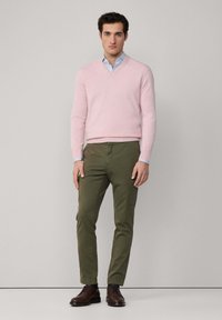 Uomo che indossa un maglione rosa chiaro a V sopra una camicia a righe blu, pantaloni cargo verde oliva e scarpe in pelle marrone scuro, in piedi davanti a uno sfondo semplice.