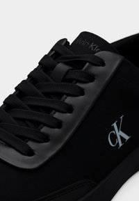 Gros plan d'une basket Calvin Klein noire avec des détails en tissu et en cuir, des lacets noirs et le logo CK sur le côté.
