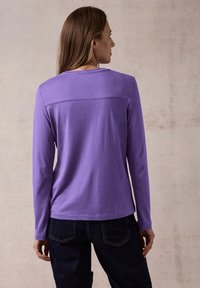 Langärmliges Oberteil aus einfarbigem lila Stoff, mit glatter Textur und geradem Schnitt, kombiniert mit dunkelblauen Jeans.
