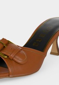 Mule en cuir marron à talon haut avec bout ouvert, boucle décorative et semelle intérieure noire portant l'inscription "GUESS" en lettres dorées.