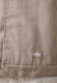 Panneau en tissu beige avec une texture côtelée diagonale. Comprend un ourlet cousu et une étiquette de marque rectangulaire dans le coin inférieur droit.