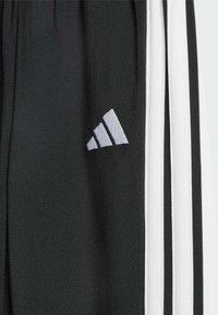 Zbliżenie czarnego materiału Adidas z białym haftowanym logo i trzema pionowymi białymi paskami po bokach.