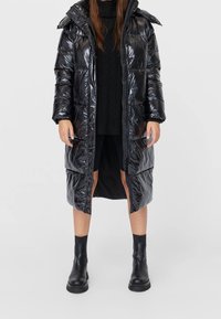 Dlouhá černá péřová bunda z lesklého materiálu s kapucí, zipovým zapínáním a elastickými manžetami. Nositá přes černý pletený svetr a šortky. Černé kotníkové boty.