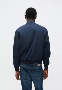 Ναυτικό bomber μπουφάν με λεία υφή, ριμπ μανσέτες και μέση, με στρογγυλή λαιμόκοψη και μινιμαλιστικό σχέδιο. Φορεμένο με μπλε τζιν.