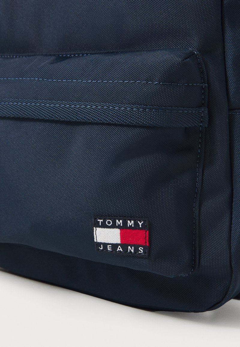 Granatowy plecak z materiału o fakturowanym wykończeniu. Posiada wyraźne logo naszywki z czerwonymi, białymi i niebieskimi akcentami. Zawiera kieszeń z przodu.
