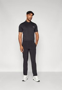 Fekete galléros póló kis logóval, karcsúsított fekete nadrág és fehér tornacipő. A modell fekete sapkát visel, egyszínű háttér előtt áll.