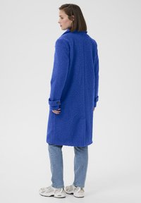 Palton pufos albastru, cu un guler structurat, mâneci lungi și manșete cu nasturi. Purtați peste blugi din denim deschis și asortați cu teniși gri.