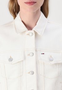 Bela denim jakna s klasičnim ovratnikom, ki ima srebrne gumbe in dva sprednja žepa. Prikazana je kontrastna šivanka in majhna logotipna podrobnost.