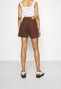Shorts de mezclilla marrones con una cintura alta, dos bolsillos traseros y una etiqueta en la cinturilla. Llevados con sandalias blancas de tiras.