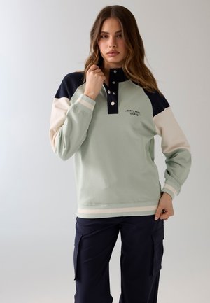 Mujer con un jersey de manga larga con bloques de color y cuello con botones, y pantalones cargo azul marino, de pie frente a un fondo liso.