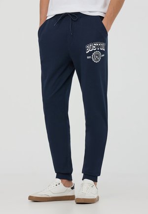 Pantaloni jogger blu navy in morbido tessuto, con vita elasticizzata e cordino, tasche laterali e una grafica bianca "Boston" sulla coscia.