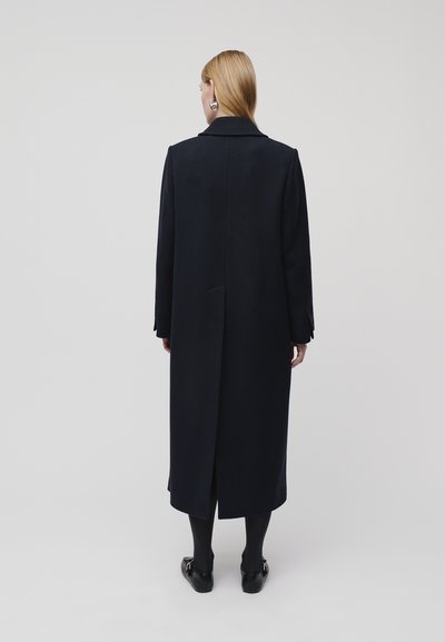 Navy long coat with a notched collar, tailored silhouette, single back vent, and smooth fabric texture, paired with black shoes.