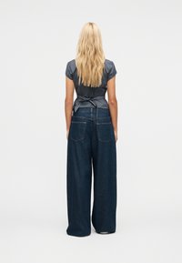 Ξανθιά γυναίκα όρθια με πλάτη, φορώντας γκρι κοντομάνικο μπλουζάκι με δεμένη μέση και φαρδιά μπλε σκούρα τζιν παντελόνια σε λευκό φόντο.