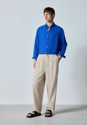 Junger Mann steht, trägt ein hellblaues Hemd mit Knöpfen, beige lockere Hose und schwarze Sandalen vor einem schlichten hellen Hintergrund.
