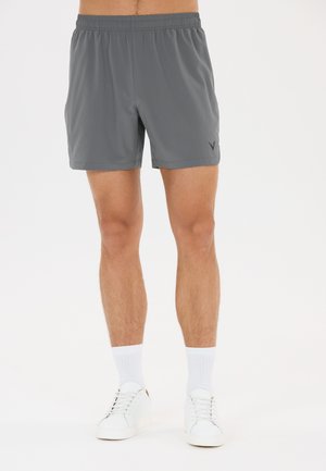 Homme portant un short de sport gris, des chaussettes blanches montantes et des baskets blanches, debout devant un fond clair uni.