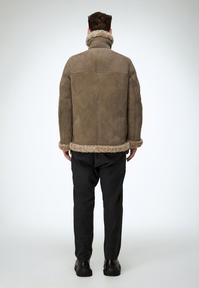 Bruine shearlingjas met een hoge kraag en zachte bontafwerking aan de manchetten en zoom, gecombineerd met zwarte broek en stevige zwarte laarzen.