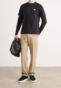 Czarny warstwowy t-shirt z małym logo, beżowe spodnie i czarne sneakersy. Trzyma czarną skórzaną kurtkę. Gładka faktura i luźny krój.