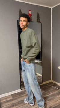 Pilkas džemperis su kišene, šviesiai mėlynos spalvos džinsai ir balti sportbačiai. Stovi šalia veidrodžio neutralaus fono sienos.