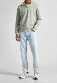 Hombre vistiendo un sudadera verde claro con cremallera de un cuarto, jeans azul claros y zapatillas blancas, de pie con una mano en el bolsillo frente a un fondo liso.