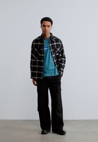 Νεαρός άνδρας στέκεται, φοράει μαύρο και άσπρο καρώ πουκάμισο πάνω από μπλε μπλούζα και μαύρο παντελόνι cargo, μπροστά σε απλό γκρι φόντο.