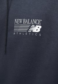 Λογότυπο New Balance Athletics με στυλιζαρισμένο "NB" και οριζόντιες γραμμές σε σκούρο ύφασμα, κοντά σε κορδόνι μαύρου φούτερ με κουκούλα.