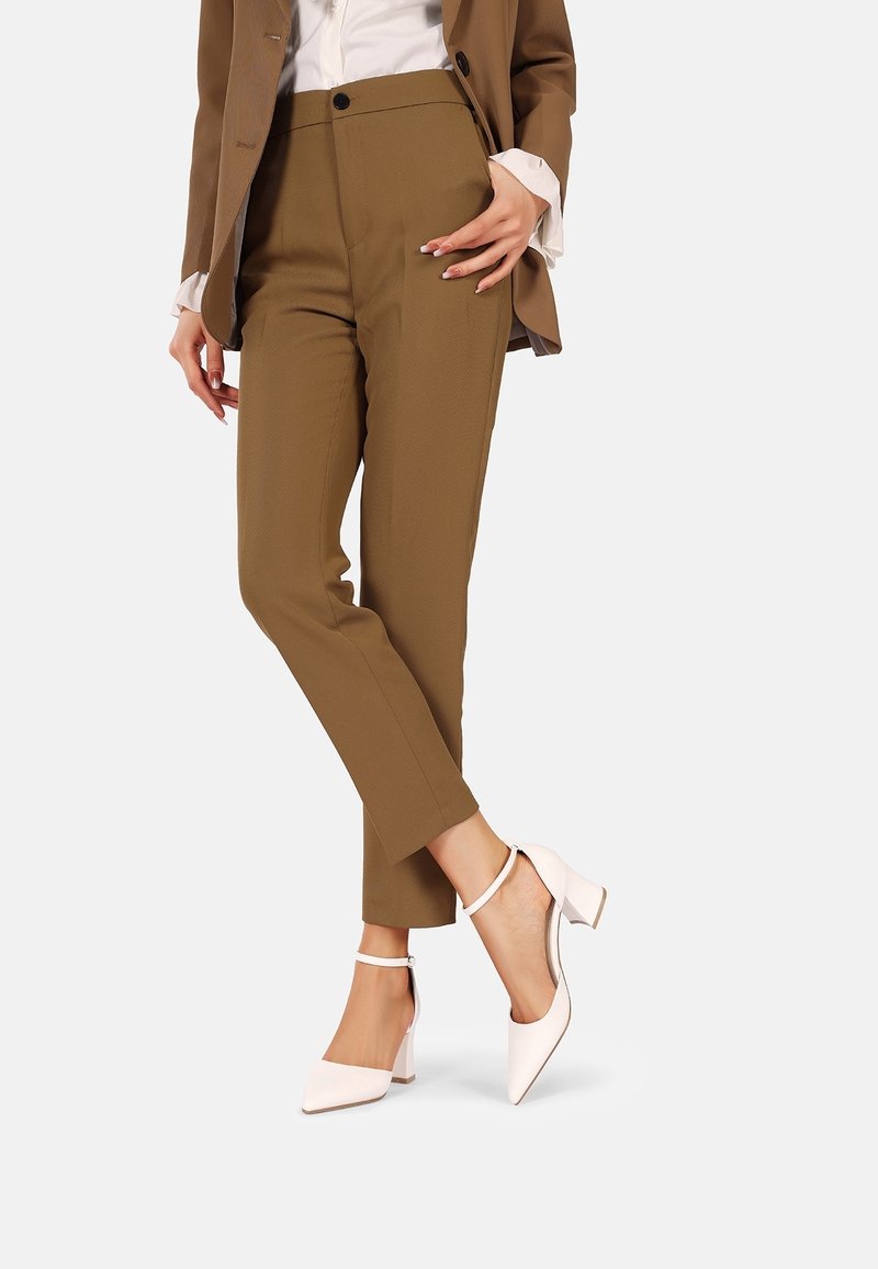 Femme portant un pantalon marron sur mesure, des talons blancs avec bride à la cheville et un blazer marron avec une chemise blanche visible aux poignets et au col.