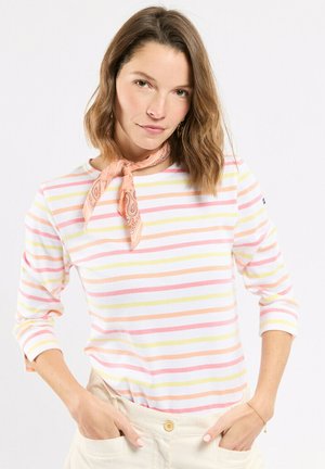 Femme aux cheveux bruns mi-longs portant une chemise à manches longues à rayures pastel, un foulard de cou à motifs pêche, et un pantalon taille haute crème, les mains dans les poches.