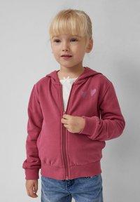 Rosa Kapuzenjacke mit weichem Texture, ausgestattet mit zwei kleinen Herzmotiven aus Glitzer auf der Brust, kombiniert mit blauen Jeans.