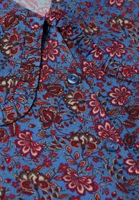 Tissu bleu avec motif floral rouge et marron, présentant des boutons bleu foncé et des fronces près du col.