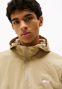 Beżowa lekka kurtka z zamkiem z przodu, kapturem i teksturowanymi akcentami. Posiada naszywkę z logo na klatce piersiowej oraz gładkie wykończenie materiału.
